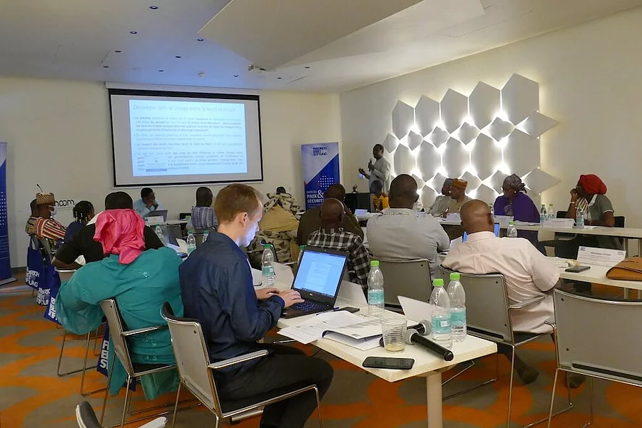 A group of young people are sitting at tables in a modern conference room during a workshop. In the foreground, a man is typing on his laptop, in the background a presentation is visible on a screen and a speaker is speaking into a microphone in front of a wall.