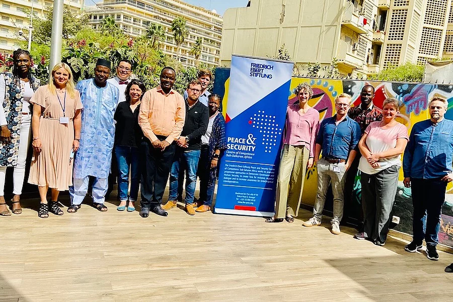 A group of around 15 people are smiling as they pose for a group photo in a sunny outdoor setting. They are standing in front of a colourfully painted graffiti wall. In the centre of the group is a large blue banner from the Friedrich Ebert Foundation bearing the words ‘Peace & Security’.