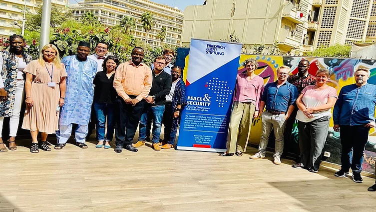 Un groupe d'une quinzaine de personnes pose en souriant pour une photo de groupe dans un espace extérieur ensoleillé. Elles se tiennent devant un mur recouvert de graffitis aux couleurs vives. Au milieu du groupe, on aperçoit une grande banderole bleue de la Fondation Friedrich Ebert portant l'inscription « Peace & Security ».